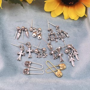 May include: A selection of silver earrings featuring cross, stud, and drop designs. Also shown are two silver safety pin earrings and two gold safety pin earrings with floral embellishments. The earrings are presented on a blue surface.
