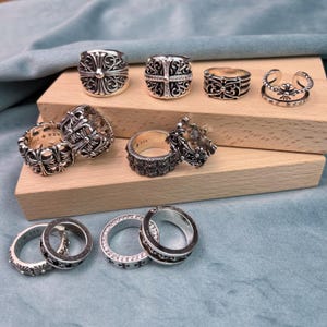 Op de afbeelding: Een verzameling zilveren ringen met verschillende ontwerpen, waaronder kruismotieven en sierlijke patronen. Sommige ringen hebben ingebedde edelstenen. De ringen worden tentoongesteld op een houten oppervlak tegen een blauwe achtergrond.