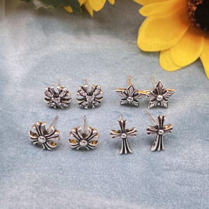 Puede incluir: Un conjunto de pendientes de plata con diseños variados: flores, estrellas y cruces. Algunos pendientes tienen pequeñas piedras preciosas transparentes. Se muestran sobre una superficie azul claro, con un girasol.