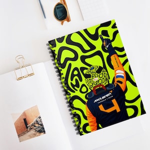 May include: Spiral-bound notebook featuring a vibrant illustration of a Formula 1 driver in a McLaren uniform. The cover has a neon green background with black abstract shapes. The driver is raising their arm in celebration.