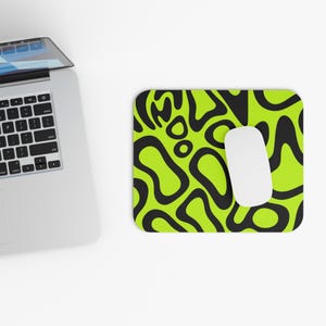 May include: A computer mouse and mousepad. The mousepad has a bright green background with a black abstract pattern. A white computer mouse is on the right side of the mousepad. A laptop is in the background.
