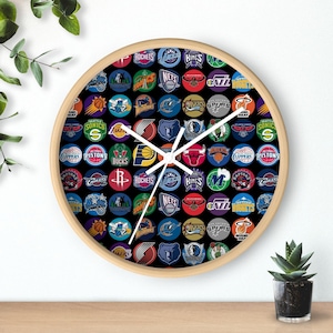 May include: A round wall clock with a light wood frame. The clock face is black and features numerous colorful circular logos of various basketball teams. White clock hands indicate the time. A small potted plant sits below the clock.