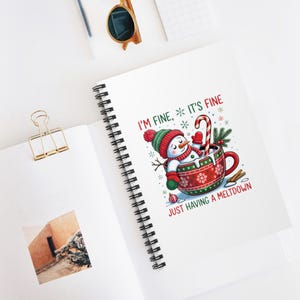 Puede incluir: Cuaderno blanco con espiral con un diseño navideño caprichoso. La ilustración representa un muñeco de nieve en una taza roja y verde con el texto "I'M FINE, IT'S FINE JUST HAVING A MELTDOWN". El cuaderno está abierto sobre una superficie blanca.