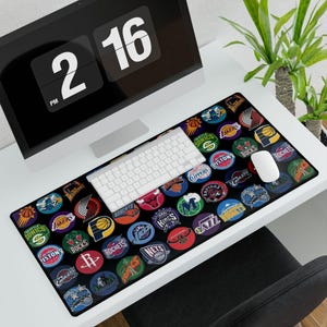 May include: Black rectangular mouse pad featuring numerous NBA team logos in a circular design. The logos are in various colors and designs, set against a black background. A white keyboard and mouse are on the pad.