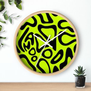 May include: A round wall clock with a light wood frame. The clock face features a vibrant neon green background with a black abstract pattern. White hands indicate the time. A small potted plant sits below the clock.