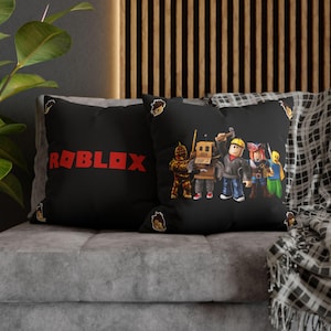 Könnte beinhalten: Schwarze Kissen mit dem Wort "ROBLOX" in Rot und einer Gruppe von Cartoon-Charakteren. Die Kissen liegen auf einem grauen Sofa, mit einer gemusterten Decke.