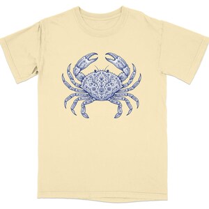 May include: A light yellow t-shirt featuring a detailed blue crab design. The crab illustration has intricate patterns and is centered on the shirt. The shirt has short sleeves and a crew neck.