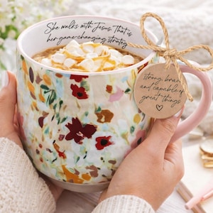 May include: A floral-patterned coffee mug with a pink handle, filled with a beverage topped with marshmallows and caramel. The mug has text that reads "She walks with Jesus. That is her game changing strategy." A tag reads "strong, brave and capable of great things."