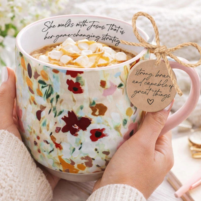 May include: A floral-patterned coffee mug with a pink handle, filled with a beverage topped with marshmallows and caramel. The mug has text that reads "She walks with Jesus. That is her game changing strategy." A tag reads "strong, brave and capable of great things."