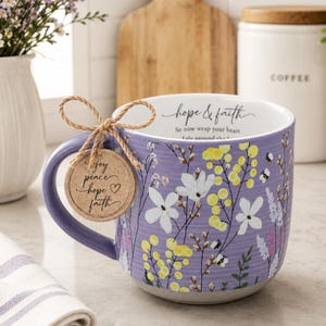 May include: A lavender ceramic mug featuring a floral pattern of yellow and white flowers with small bees. The interior rim displays the text hope & faith. A round cardboard tag tied to the handle with twine reads joy, peace, hope, faith.