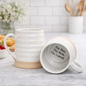May include: Two white ceramic mugs with a beige base and handle. One mug is upright, the other is on its side, revealing the text "The Man. The Myth. The Legend." inside. The mugs have a ribbed design.