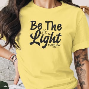 May include: A pale yellow short-sleeved t-shirt featuring the black text "Be The Light" with a small lightbulb icon replacing the letter "i" in "Light" and the scripture reference "Matthew 5:14" printed underneath in a smaller font.