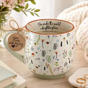 May include: A white ceramic mug featuring a colorful wildflower pattern, a brown rim, and a light green handle. The interior rim displays the text You make the world a brighter place Philippians 1:3. A brown tag tied with twine reads You are amazing.
