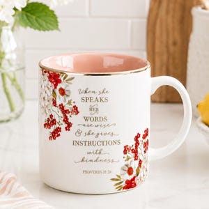 May include: A white ceramic mug with a soft pink interior and gold rim. The exterior features a floral design with red and white flowers and gold text that reads: When she SPEAKS HER WORDS are wise & she gives INSTRUCTIONS with kindness PROVERBS 31:26.