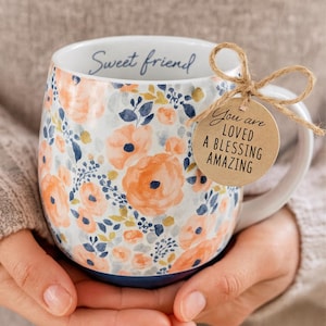 May include: White ceramic mug with a floral pattern in orange and blue. The mug has the words "Sweet friend" on the rim and a tag that says "You are LOVED A BLESSING AMAZING".