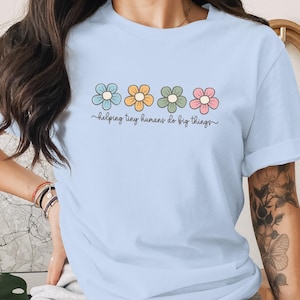 May include: A light blue short-sleeve t-shirt featuring a graphic of four stylised flowers in light blue, yellow, sage green, and pink. Below the flowers, the text "helping tiny humans do big things" is printed in a dark brown cursive script.