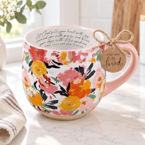 May include: A ceramic mug featuring a vibrant floral pattern in shades of orange, pink, and yellow on a white background. The interior rim displays the text Let God fill your heart with joy, surround you with peace, and bless your days with His goodness. ROM. 15:13 AND NUM. 6:24-26. A brown paper tag tied to the handle reads YOU ARE loved.