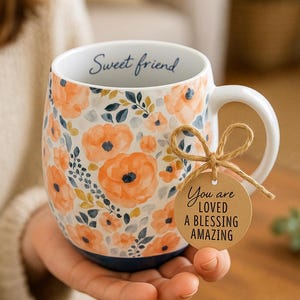 May include: White ceramic mug with orange floral pattern and the words "Sweet friend" on the rim. A brown tag tied to the handle reads "You are LOVED A BLESSING AMAZING". The mug is held in a person's hand.