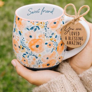 May include: A floral ceramic mug with a white handle and a blue base. The mug features orange and blue flower patterns and the words "Sweet friend" on the rim. A tag tied to the handle reads "You are loved, a blessing, amazing."