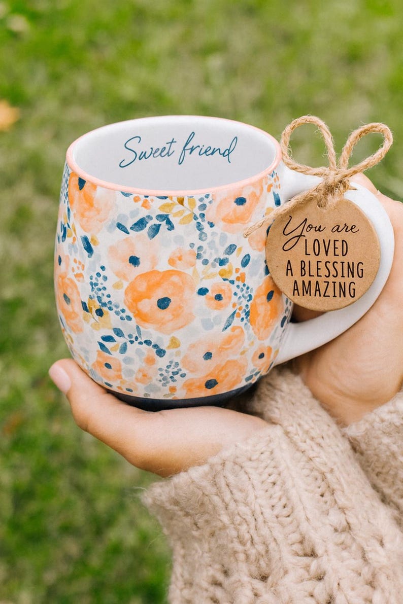 May include: A floral ceramic mug with a white handle and a blue base. The mug features orange and blue flower patterns and the words "Sweet friend" on the rim. A tag tied to the handle reads "You are loved, a blessing, amazing."