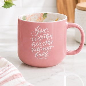 May include: A pink ceramic mug featuring white cursive text that reads God is within her, She will not fall, PSALM 46:5. The interior of the mug is decorated with a soft floral pattern in shades of peach, pink, and green, visible from the top rim.