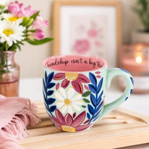 May include: A light green ceramic mug featuring a painted floral pattern with white, yellow, and dark pink flowers and blue leaves. The interior of the mug is pink and displays the black text, Friendship isn't a big thing.