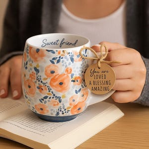 May include: White ceramic mug with orange floral design and blue accents. The mug has the words "Sweet friend" inside and a tag that says "You are loved a blessing amazing". The mug is sitting on an open book.