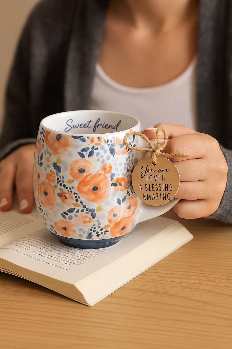 May include: White ceramic mug with orange floral design and blue accents. The mug has the words "Sweet friend" inside and a tag that says "You are loved a blessing amazing". The mug is sitting on an open book.