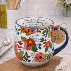 May include: Floral ceramic mug with a blue handle and rim, featuring a design of red roses, green leaves, and blue berries on a white background. The mug has the text "Confidently trust God is with you..." and a tag that says "You are loved".