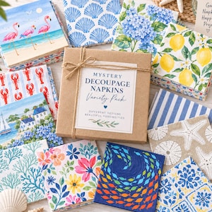 May include: A brown cardboard box tied with twine, labeled Mystery Decoupage Napkins Variety Pack, surrounded by stacks of paper napkins featuring various prints including flamingos, seashells, blue hydrangeas, lemons, lobsters, and fish.