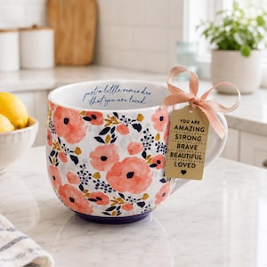 May include: A large white ceramic mug with a floral pattern in coral and navy blue. The mug has a purple base and a tag tied with a peach ribbon that reads "YOU ARE AMAZING STRONG BRAVE BEAUTIFUL LOVED". The mug also has the text "just a little reminder that you are loved".
