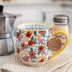 May include: Floral patterned ceramic mug with a yellow handle and rim. The mug features red, pink, and green floral designs on a white background. A small tag that says "YOU ARE loved" is attached to the handle. The mug is sitting on a beige patterned napkin.