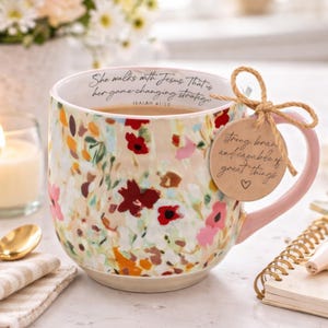 May include: Floral-patterned ceramic mug with a pink handle. The mug features a cream-colored base with a colorful floral design in red, pink, and orange. Text on the mug reads "She walks with Jesus. That is her game-changing strategy." A tag reads "strong, brave and capable of great things."