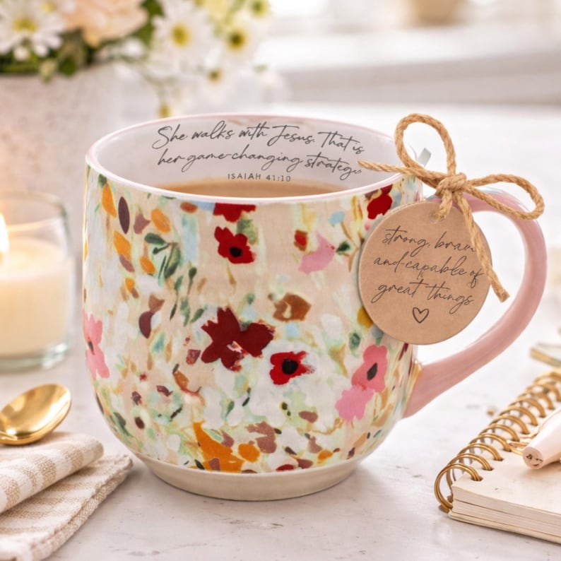 May include: Floral-patterned ceramic mug with a pink handle. The mug features a cream-colored base with a colorful floral design in red, pink, and orange. Text on the mug reads "She walks with Jesus. That is her game-changing strategy." A tag reads "strong, brave and capable of great things."