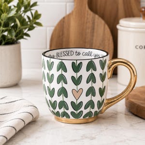 May include: A white ceramic mug featuring a repeating vertical pattern of green heart-shaped leaves and one light pink heart. The mug has a metallic gold handle and base, with the text "BLESSED to call you" printed along the inner rim.