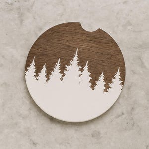 May include: A round car coaster with a forest silhouette design. The top half is a dark brown wood grain, and the bottom half features a white forest silhouette. The coaster has a small notch for easy removal from a car cup holder.