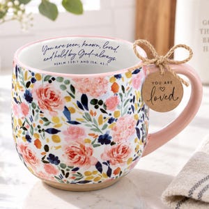 May include: A white ceramic mug featuring a colorful floral pattern of pink roses, blue leaves, and yellow accents. The interior rim displays the text: You are seen, known, loved, and held by God-always. Psalm 139:1 and Isa. 43:1. A brown tag reads: You are loved.