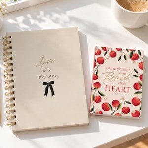 May include: A spiral-bound notebook with a cream cover featuring the text love who you are and a black bow icon, alongside a smaller book titled Mini Devotions to Refresh Your Heart with a red cherry and white blossom pattern.
