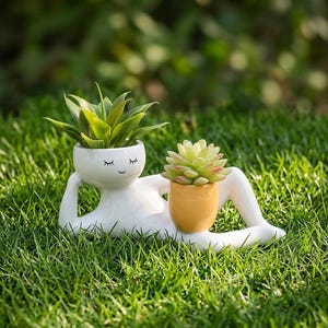 May include: A white ceramic planter shaped like a reclining figure with a serene face, closed eyes, and a small smile. The figure holds a green succulent in its head and a separate, smaller yellow pot containing a succulent between its legs.