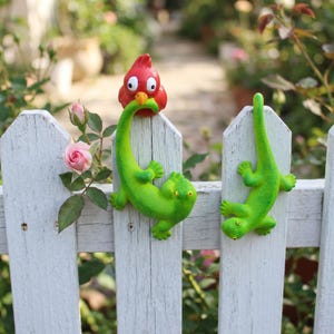 May include: Two bright green lizard figurines and one red bird figurine perched on a white wooden picket fence. The lizards have textured skin and yellow eyes, while the red bird has large white eyes and a yellow beak, all mounted on the fence.