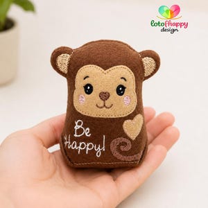 Puede incluir: Un pequeño peluche de mono hecho de fieltro marrón con cara color canela, ojos negros bordados, mejillas rosadas y nariz marrón. El frente presenta el texto bordado Be Happy! en blanco, un corazón color canela y un diseño de cola en espiral.