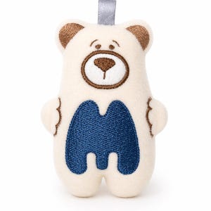 Pode incluir: Um brinquedo de pelúcia em forma de urso creme, com detalhes bordados em marrom nas orelhas, olhos e nariz. O urso tem uma letra "M" azul bordada na barriga e uma fita cinza na parte superior.