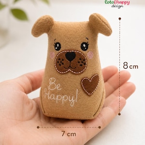 May include: A tan felt dog plushie featuring embroidered black eyes, a dark brown muzzle, pink cheeks, and a brown heart patch. The text "Be Happy!" is embroidered in white. Dimensions are 8 cm in height and 7 cm in width.
