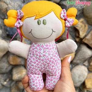以下が含まれることがあります： 明るい黄色の髪を2つのおさげにし、ピンクと白の花柄の小さなリボンで結んだ布製のドール。刺繍された緑色の目とステッチされた口元が特徴で、ピンクの花柄があしらわれた白いロンパースを着用しています。