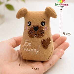 May include: A tan felt dog plushie featuring embroidered black eyes, a dark brown muzzle, pink cheeks, and a brown heart patch. The text "Be Happy!" is stitched in white thread. The item measures 9 cm in height and 7 cm in width as indicated.