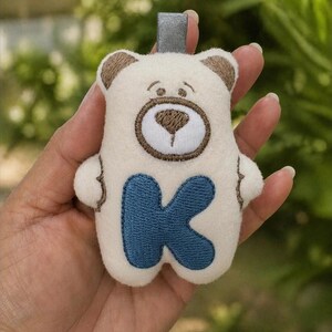 Pode incluir: Um bicho de pelúcia em forma de urso creme com detalhes marrons nas orelhas e focinho, e a letra "K" azul na barriga. O bicho de pelúcia tem uma alça cinza na parte superior para pendurar.