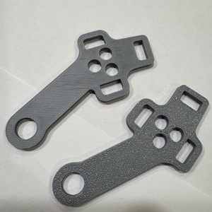 May include: Two grey, 3D-printed brackets. Each bracket has a long, rectangular arm with a circular hole at the end. The other end features a square shape with three circular holes and two rectangular openings.