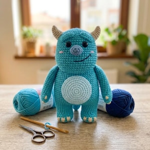 May include: A turquoise crochet monster doll with a light blue belly patch, two small beige horns, and black plastic eyes. The doll is positioned between two skeins of blue yarn, with a wooden crochet hook and metal scissors placed in front.
