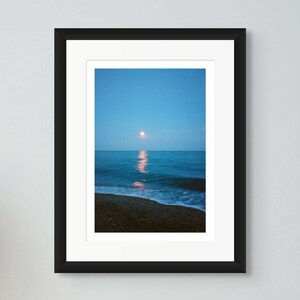May include: A framed photograph of a seascape, a black camera, and a vase of flowers on two wooden stools. The photograph shows a moonlit ocean scene. The vase is light green and tan.