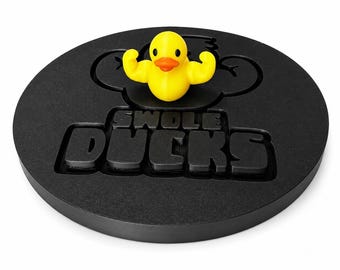 May include: A circular black base featuring the embossed text SWOLE DUCKS in a bold font. A small yellow rubber duck with an orange beak and muscular arms is attached to the top center of the base, positioned above the text.